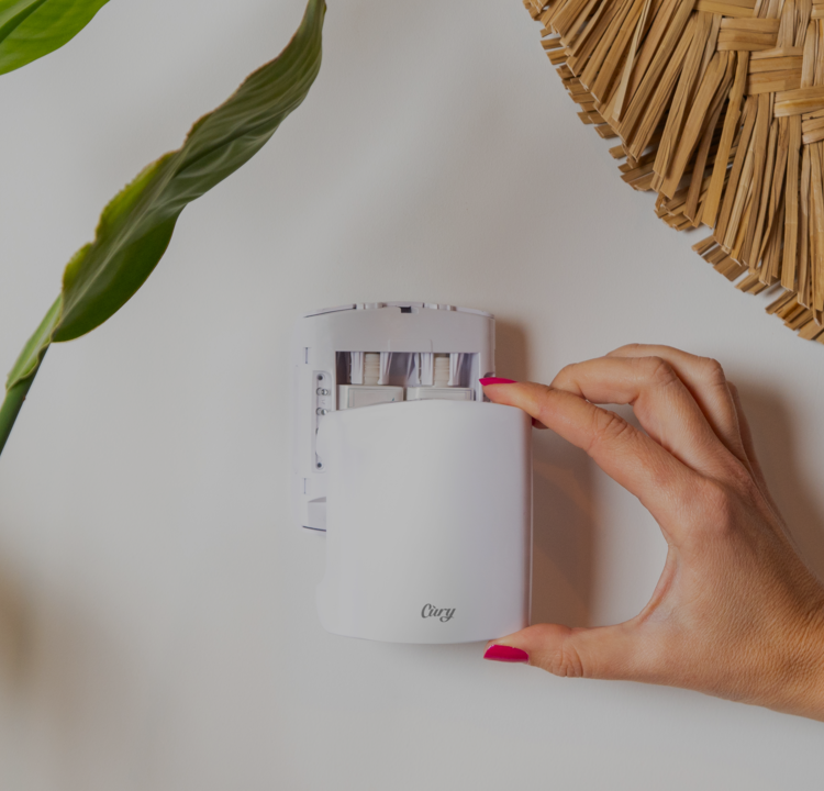 A hand with pink nail polish adjusts a white Clary device on a wall, surrounded by a green leaf and part of a woven basket.