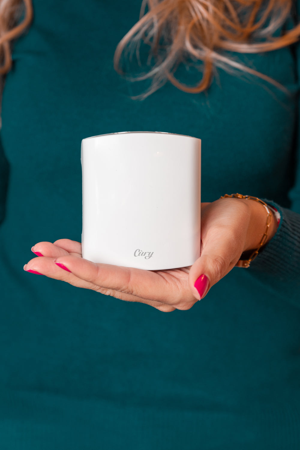 A person with long hair and pink nail polish holds a small, white cylindrical device labeled Cudy against a teal background.