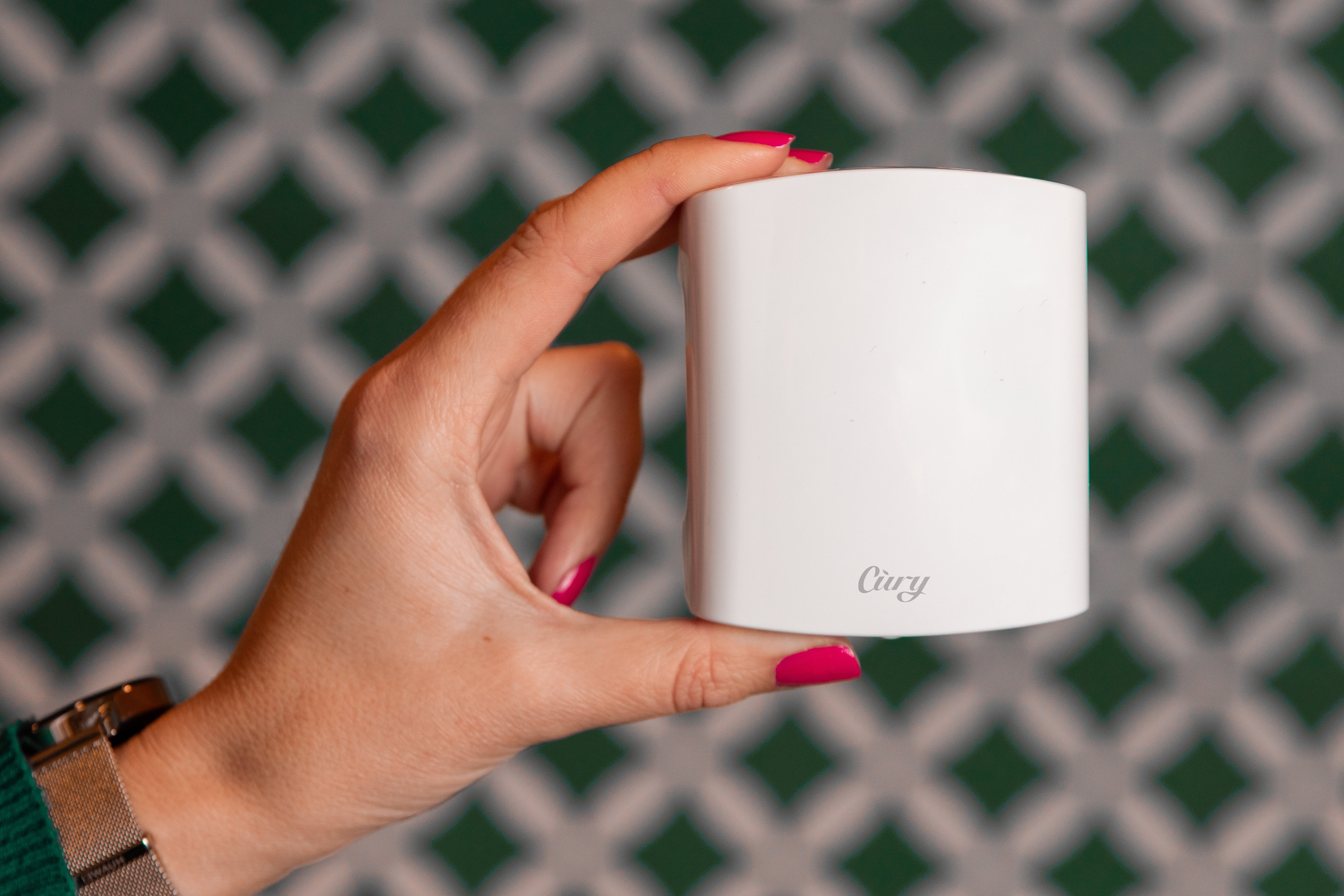 A hand with pink nail polish holds a white mug labeled “airy” against a green and white patterned background.