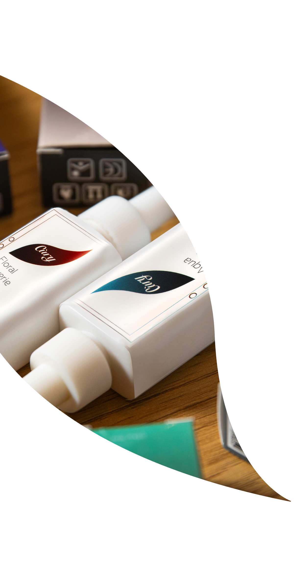 Two white rectangular bottles with colored labels lying on a wooden surface, partially visible through a curved, leaf-shaped frame; additional packaging is in the blurred background.