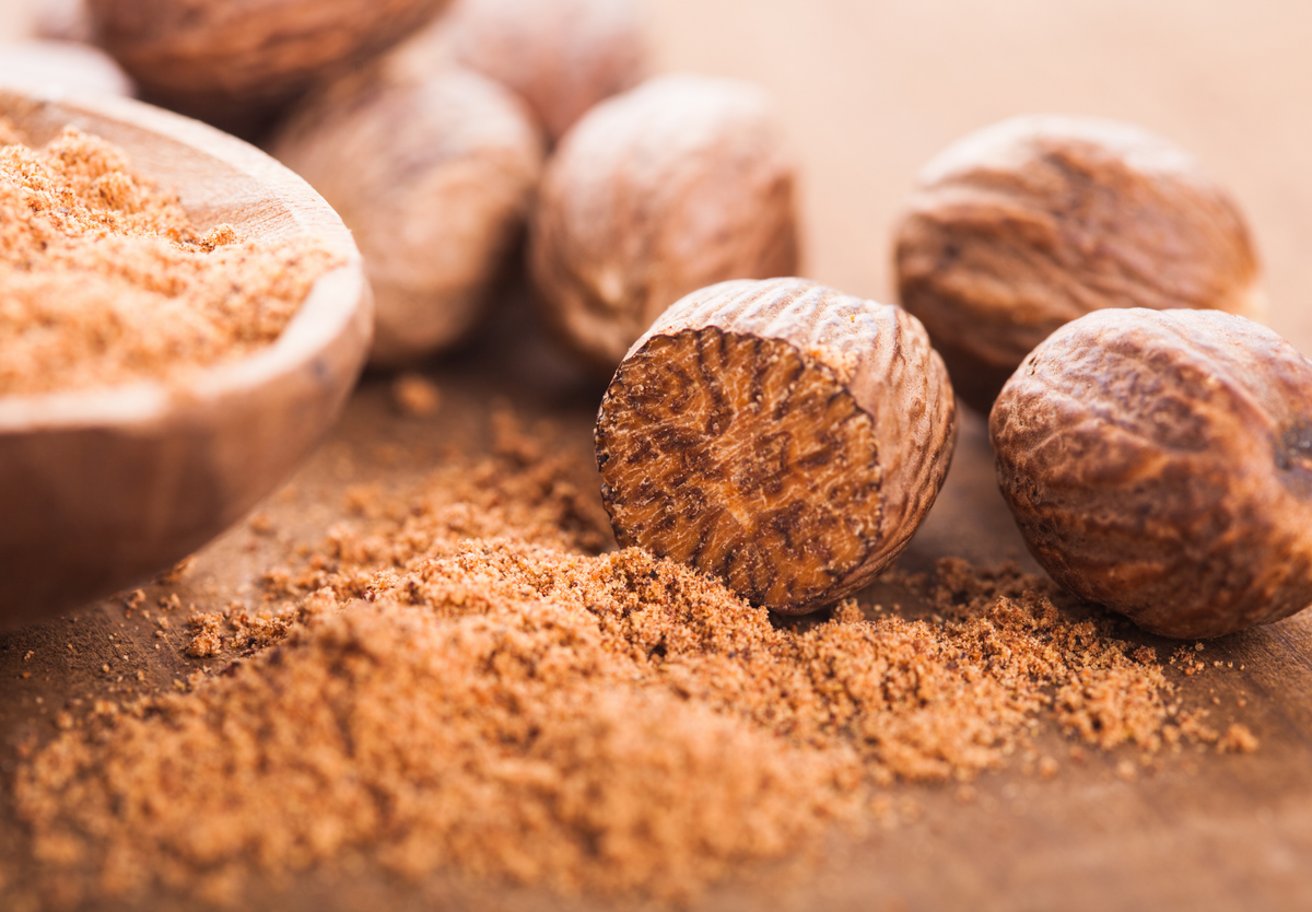 Whole nutmeg and ground nutmeg are shown on a wooden surface. A cut nutmeg seed is in focus, with its textured interior and scattered brown spice powder nearby. Other whole nutmegs are blurred in the background.