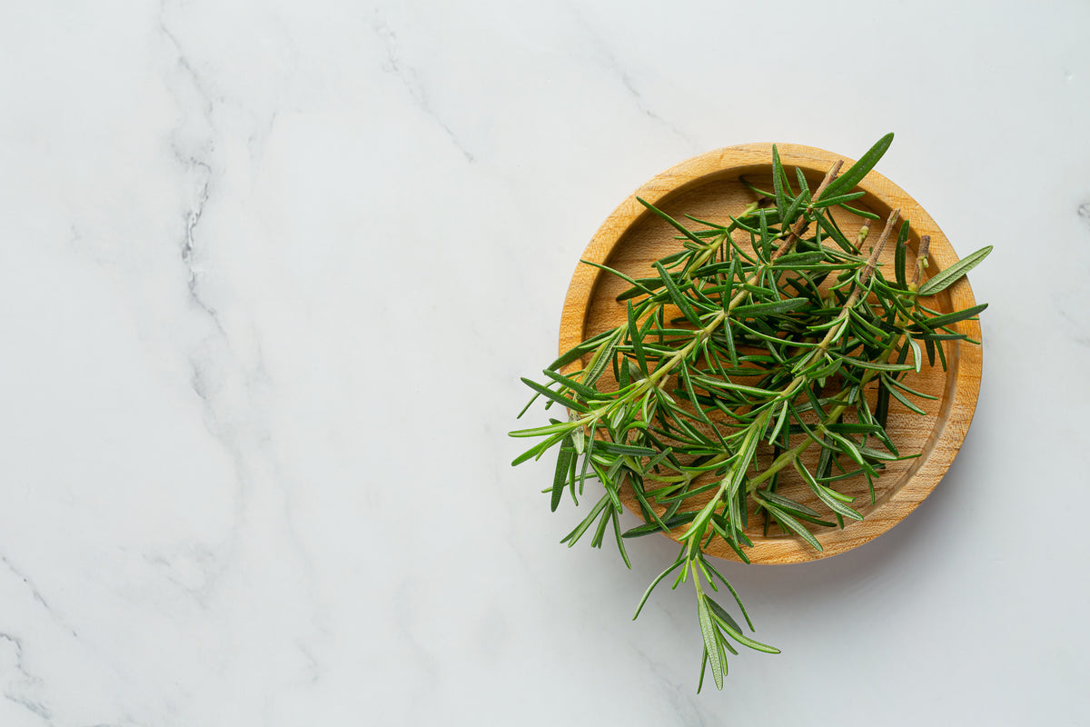 A small wooden bowl filled with fresh rosemary sprigs sits on a white marble surface.