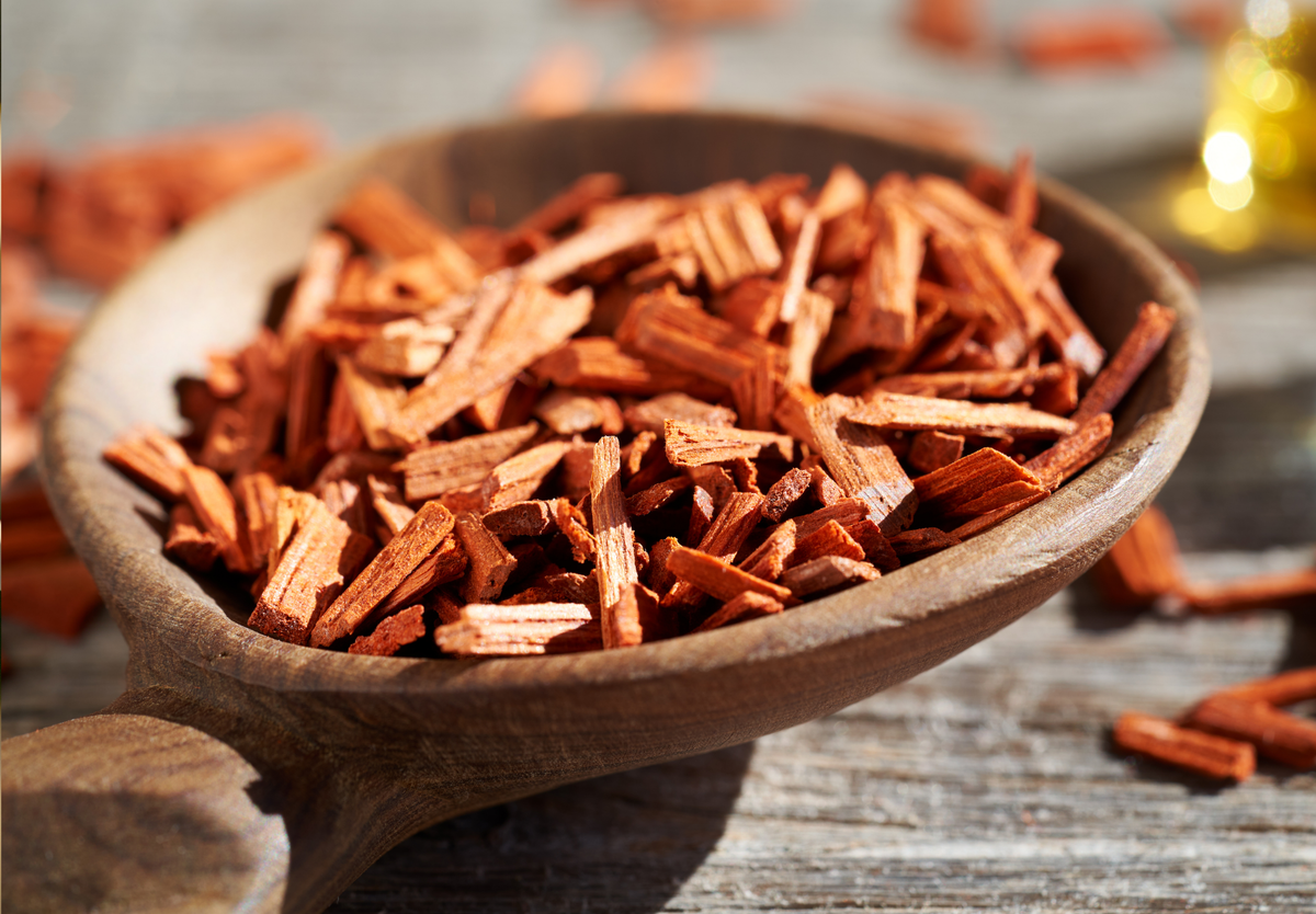 A close-up of reddish-brown sandalwood chips in a wooden spoon, resting on a rustic wooden surface.